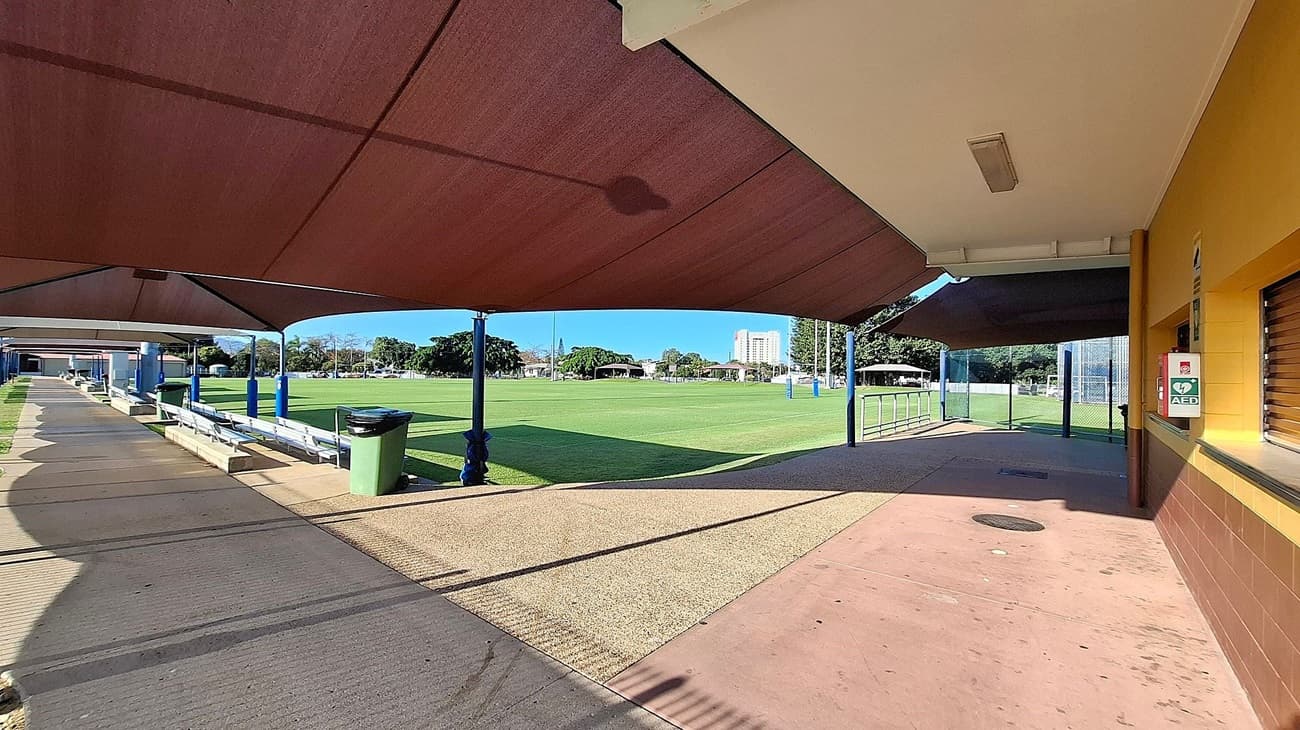 A sports field with a well-maintained green lawn is in the centre. On the left, there is a covered walkway with benches and a green bin. The right side features a building with a yellow and brown exterior, and an AED device mounted on the wall. The ceiling is covered with brown shade sails supported by blue poles. In the background, there are trees and a tall building visible.