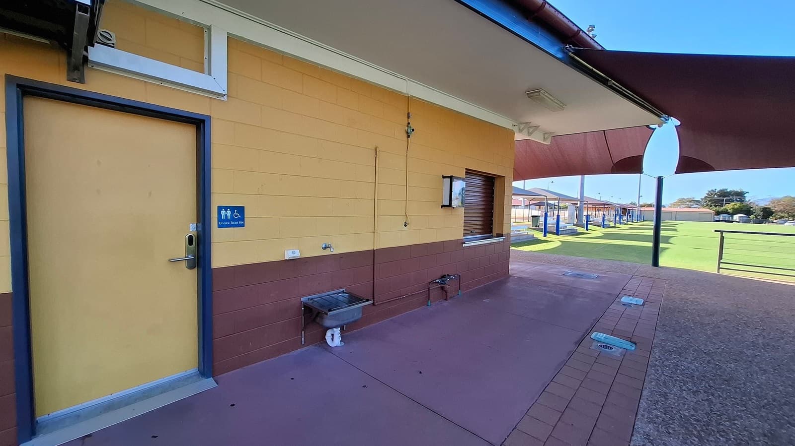 A building with a yellow and brown exterior. On the left, there's a door with a sign indicating a unisex and accessible toilet. Below the sign is a small sink with a tap. To the right, a service window is closed with a shutter. The ground is paved with a mix of purple and brown tiles. In the background, a grassy field is visible, along with covered seating areas and a railing. The ceiling has a large shade structure extending outward.