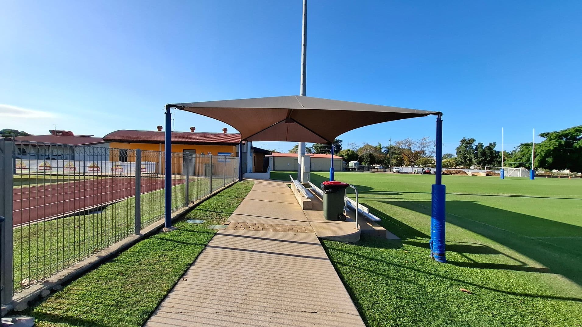 A sports field with a covered seating area in the centre, featuring a triangular shade structure supported by blue poles. To the left, a wire fence runs alongside a red track, with a yellow building in the background. On the right, a green field with rugby posts and trees in the distance. A pathway leads to the seating area, with a rubbish bin nearby. The ground is a mix of grass and concrete.