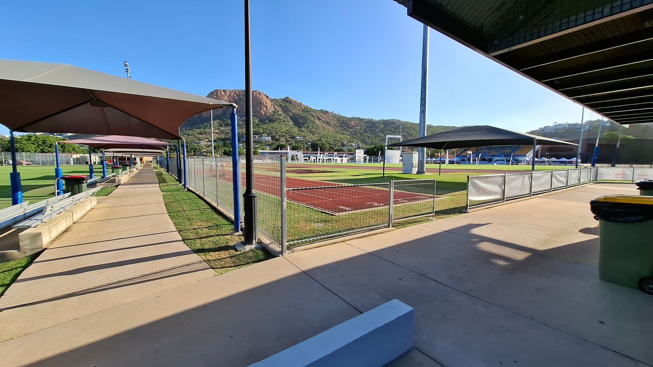 A sports facility with a covered walkway on the left, featuring blue poles and benches. The walkway is lined with grass and leads to a building in the distance. On the right, there is a fenced athletic field with a red track and green grass. A large mountain is visible in the background. The foreground has a concrete path with a green rubbish bin. The ceiling of the covered area is visible on the right.