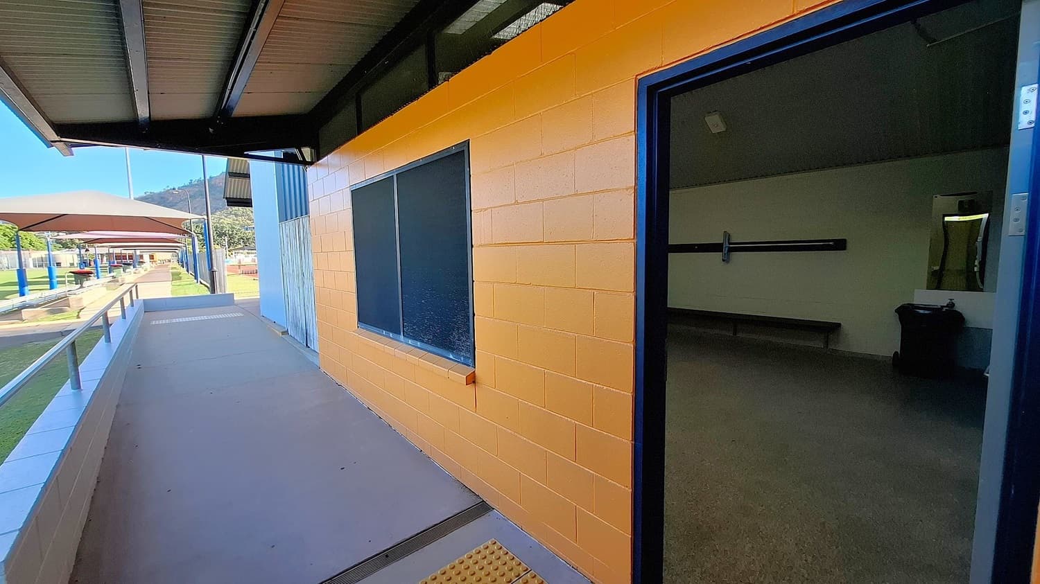 A walkway with a metal railing on the left leads to a building with a yellow brick wall on the right. The wall has a dark window and an open door leading to an interior room with a bench and a bin. The ceiling is metal, and there are covered outdoor areas with blue pillars in the background. A mountain is visible in the distance.