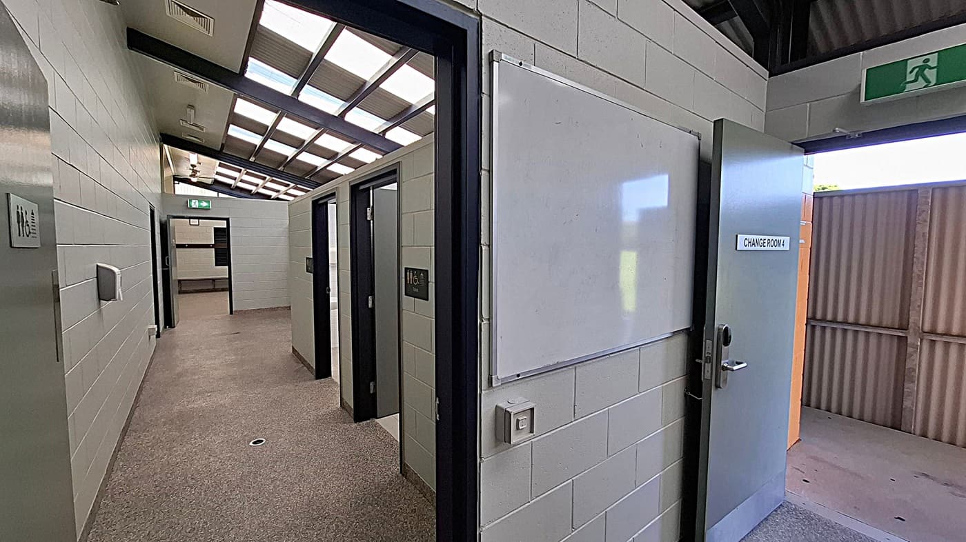 A corridor with light grey brick walls and a beige carpeted floor. On the right, a door marked "CHANGE ROOM" is open, leading outside. A whiteboard is mounted on the wall beside it. On the left, there are several doors, one with a sign indicating a toilet. The ceiling features a series of skylights, allowing natural light to illuminate the space. An exit sign is visible above one of the doors.