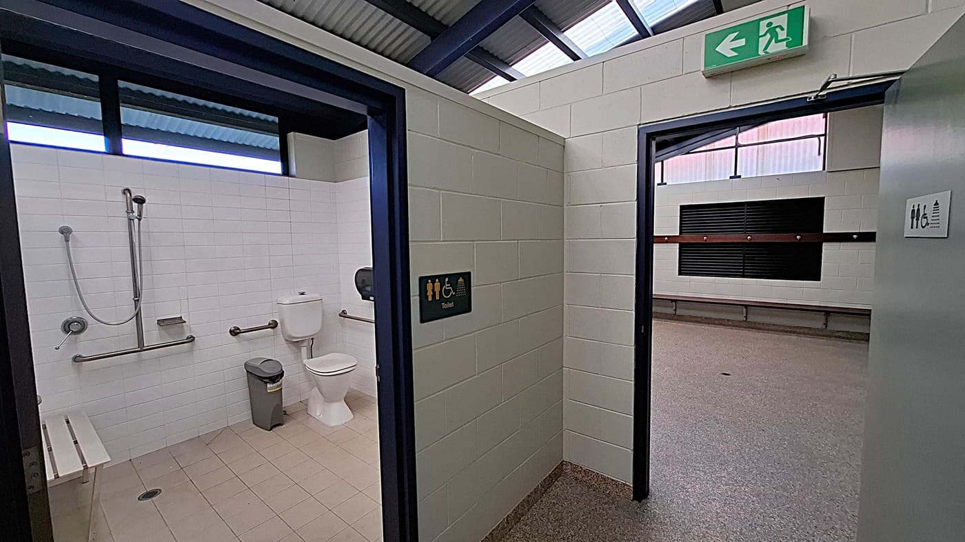 A public restroom with a white-tiled accessible toilet on the left, featuring a shower, grab bars, a toilet, and a bin. The floor is light brown tile. To the right, an open door leads to a larger room with grey flooring and a bench along the wall. Above the door is an emergency exit sign. The ceiling has exposed beams and translucent panels.