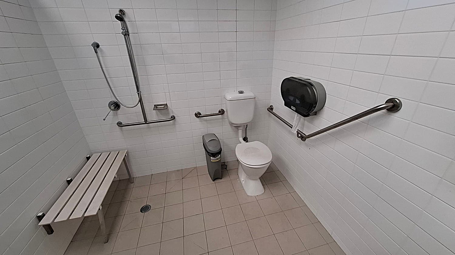 A bathroom with white tiled walls and a beige tiled floor. On the left, there is a shower area with a handheld showerhead and a metal bench. In the centre, a toilet with a white cistern is positioned next to a grey bin. On the right, a large toilet paper dispenser is mounted on the wall. Metal grab bars are installed around the toilet and shower area for accessibility.