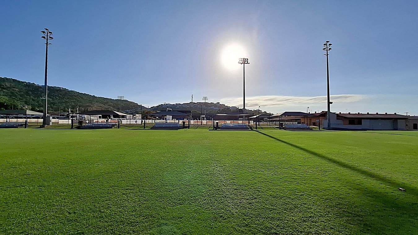 A grassy sports field with two tall floodlights on either side. In the background, there are low buildings and a hill covered with trees. The sun is positioned high in the sky, casting long shadows across the field. The grass is well-maintained, and there are a few benches near the fence that surrounds the field.