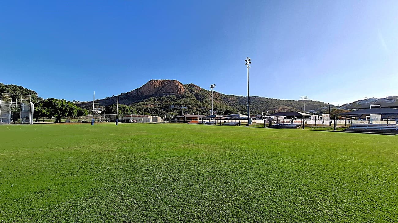 A sports field with green grass in the foreground. On the left, there are tall netted structures and trees. In the centre, rugby posts are visible. To the right, there are several buildings and seating areas. In the background, a large rocky hill with vegetation rises against a clear blue sky. Floodlights are scattered around the field.