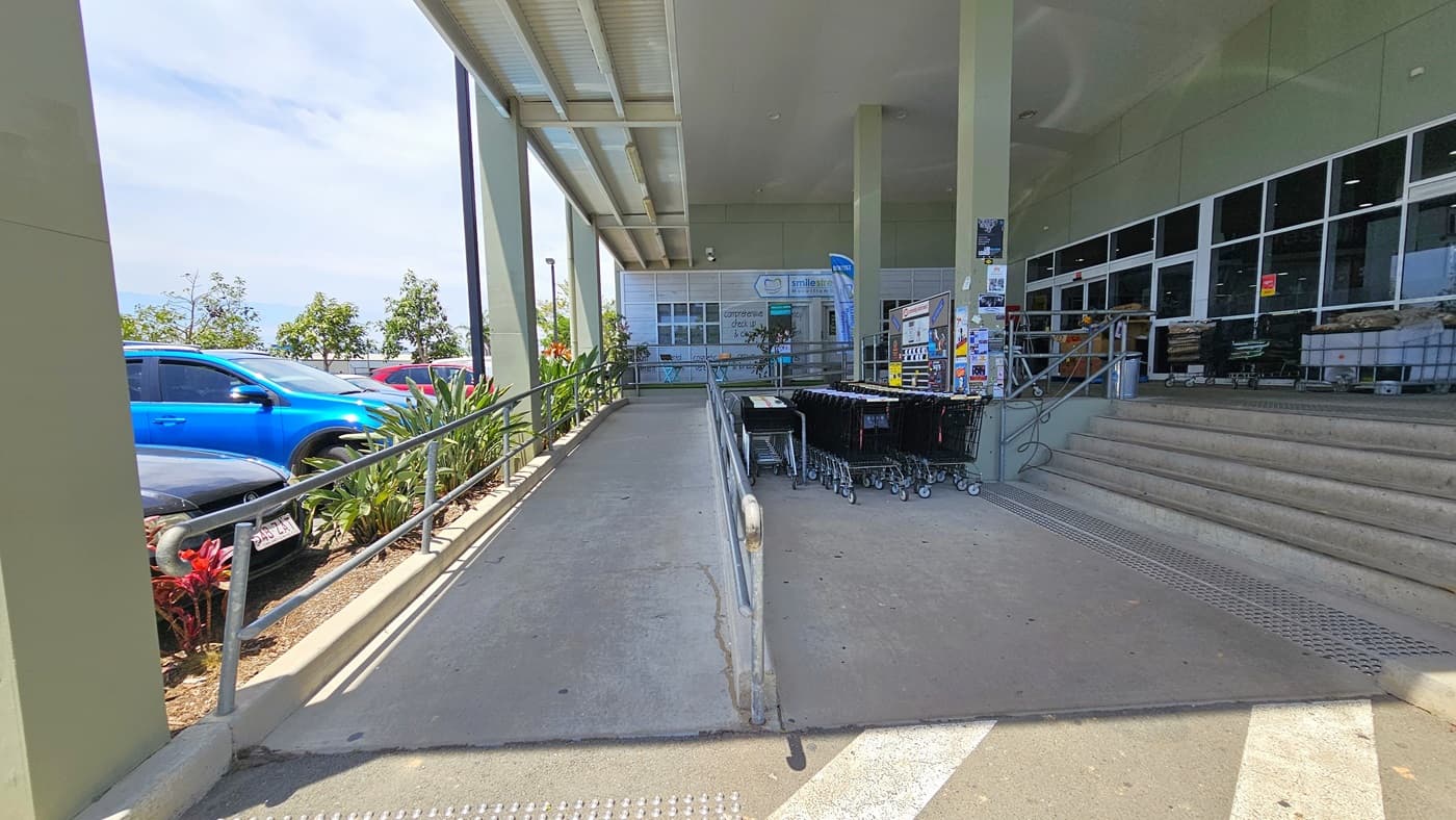 A closer view of the ramped and 5-step staircase entrances both with truncated domes before entering. Safety handrails are placed at both entrances. Adjacent to the shopping trolleys, posters and signage are placed on a nearby wall. Going to the ramped entry leads to where “smile street” is situated.