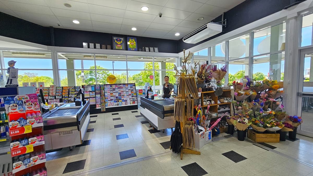Another angle of the checkout zone. Two checkout counters are apparent, where on the right counter a female checkout staff member is prominent. On the right, display racks filled with greeting cards, gift wraps, umbrellas, and magazines are situated. There’s also a colorful selection of fresh flower bouquets near the glass door. On the top of the middle area, two framed jerseys alongside several frames are apparent.