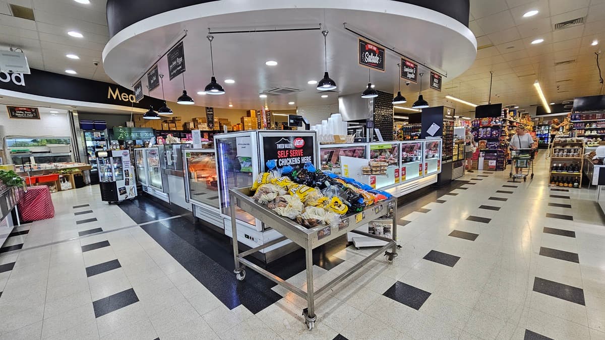 A closer look at the Delicatessen section. “Self-serve chickens” are visibly placed in the center of the Delicatessen near a steel cart loaded with products. Consumers are apparent on the right side corner adjacent to the three-layer shelves. Fluorescent lights and banners overcome the ceiling of the Delicatessen section. Striking black tiles conquer both sides of the area as well.