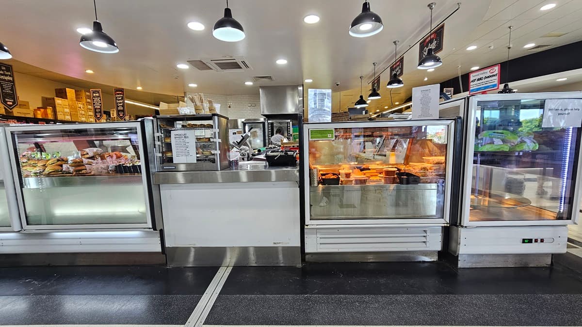 A closer look at the front view of the Delicatessen. Rows of fridges filled with burgers, cooked chicken, and other delicacies are prominent. Names of the delicacies are evidently placed above the fridges on the right. An employee is evident at the far end. Used and unused boxes are noticeable above the shelves on the far left.