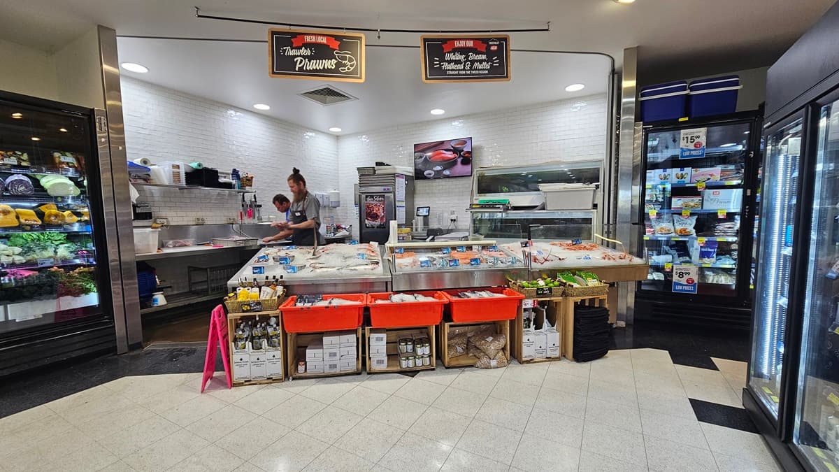 Seafood and meat section. Two banners are noticeably hanging on a black steel rod above the ceiling. Shop assistants are apparent, as well as a wall decor inside the seafood and meat section. Frozen prawns and fish are visibly displayed and underneath are beverages and other products. On the right, two fridges are prominent.