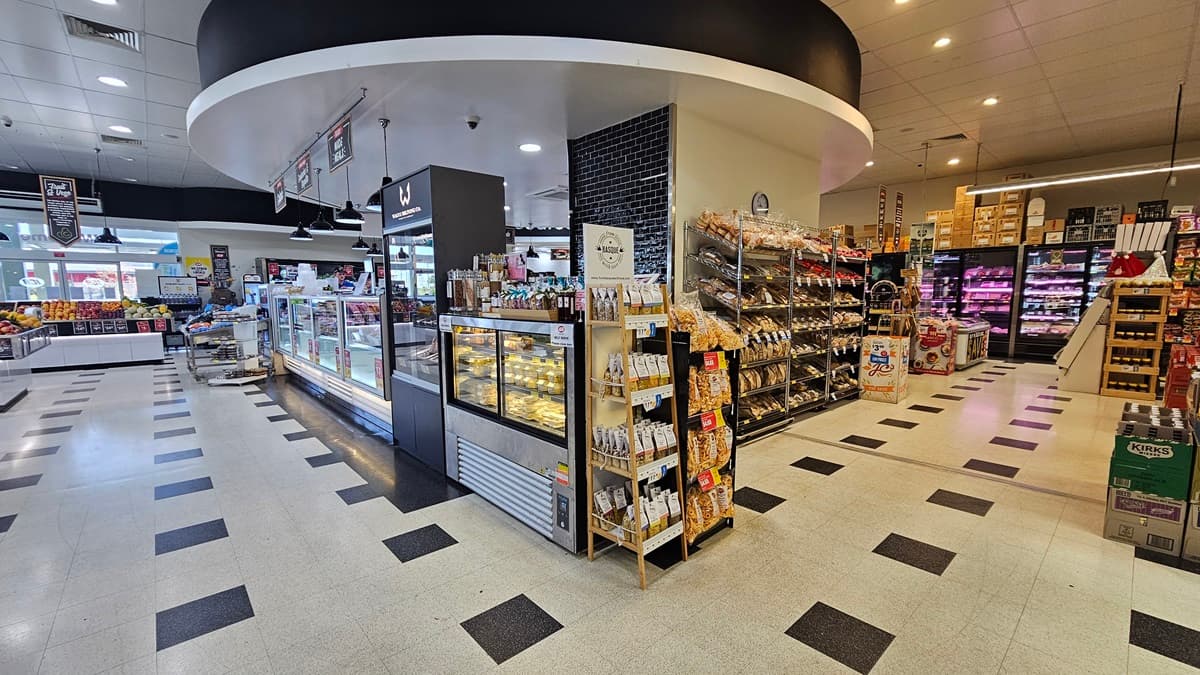Rear view of the Delicatessen section. Multi-layer racks filled with grocery items such as chips, sweets, bottled drinks, and bread. The intricate circular design in black and white color of the Delicatessen ceiling is noticeable. The pathways on each side are spacious. On the far right, fridges filled with milk, cheese, and other dairy products are neatly organized with several boxes on top.