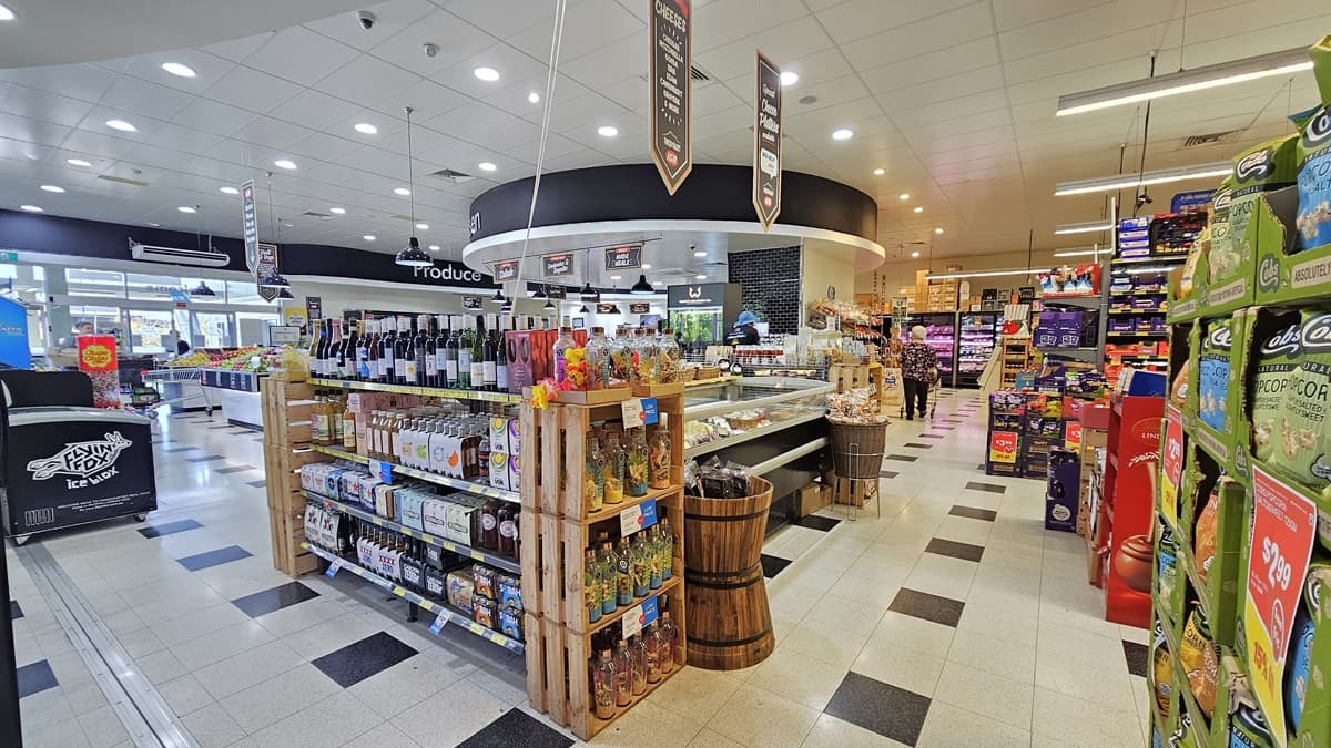 A closer look at a Grocery Aisle inside the Tweed Valley IGA. A three-layer shelf filled with wines and liquors is visible near the wooden buckets and the displayed delicacies. On the right, groceries like popcorn, sweets, and chips are neatly organized. On the left, a black freezer with “flying fox” written in it is noticeable.