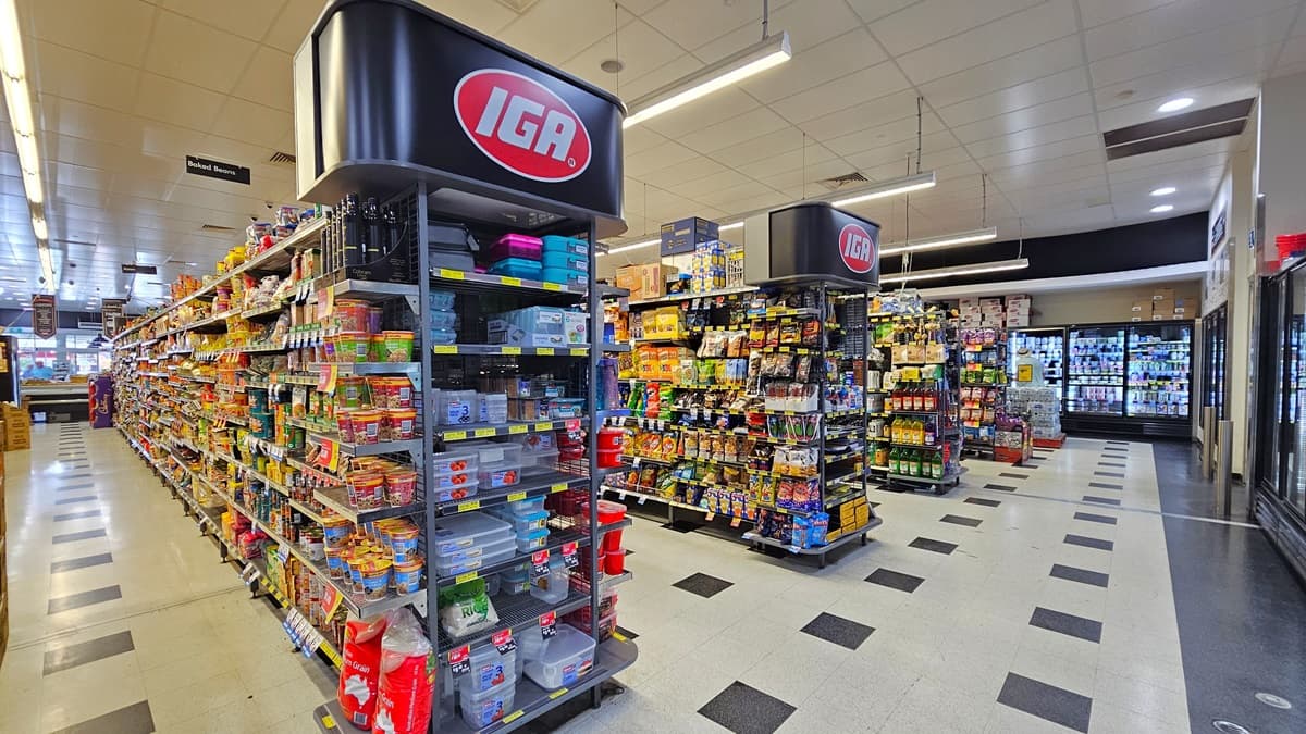 A closer look at the grocery aisles. Four multi-layer racks are apparent with sweets, chips, liquors, and tupperwares. Above the two racks are the logo and name of the building, placed in an orderly manner. Fluorescent lights illuminate the grocery aisles. On the right side, rows of fridges are noticeable.
