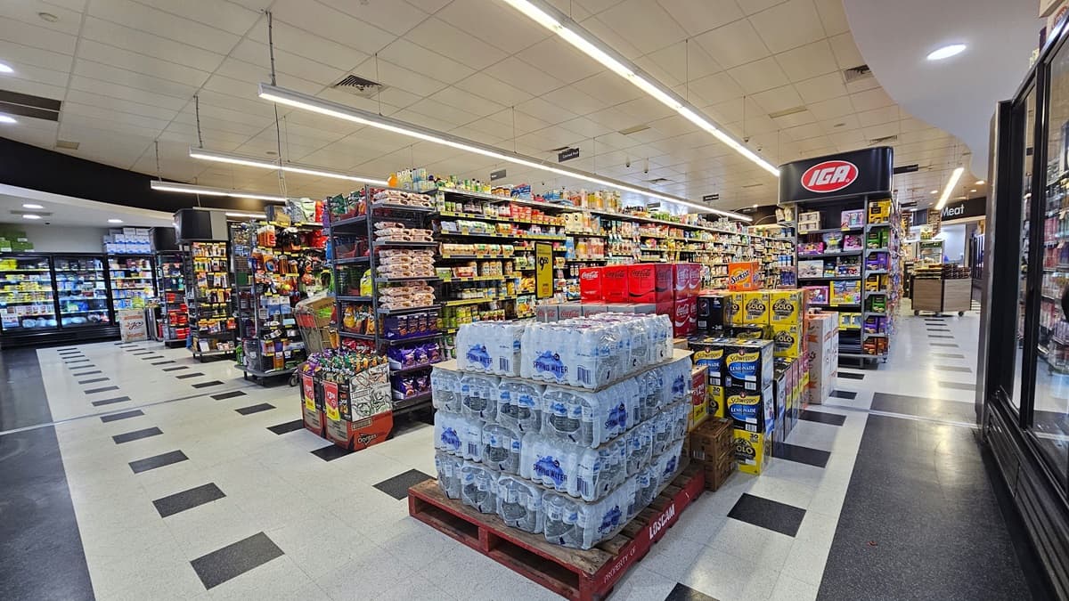 Another angle of the grocery aisles. Piles of drinking water are placed alongside other beverages. Rows of multi-layer racks filled with groceries dominate the aisle. On the far left side, rows of fridges that contain sweets are noticeable, along with the boxes that are placed on top of them.