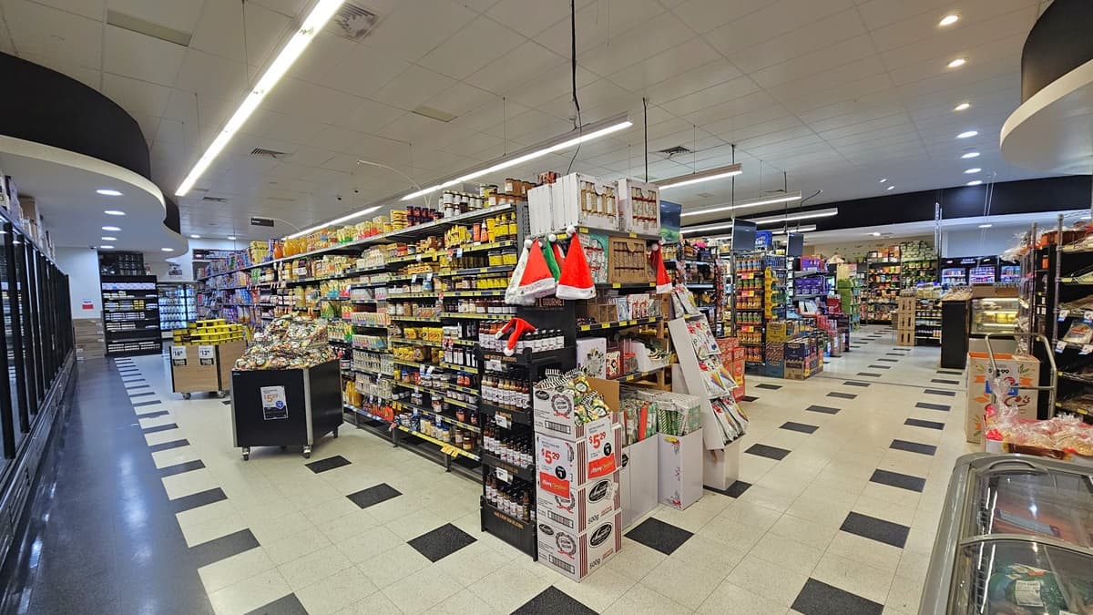 A closer look at the pathways beside the grocery aisles. Santa hats are visibly placed above layers of liquors beside the Christmas wrapping paper and other Christmas decorations. Two box trolleys with sweets and other products are apparent on the left. On the pathway on the right, a trolley with products is noticeably placed near a freezer and a multi-layer rack.
