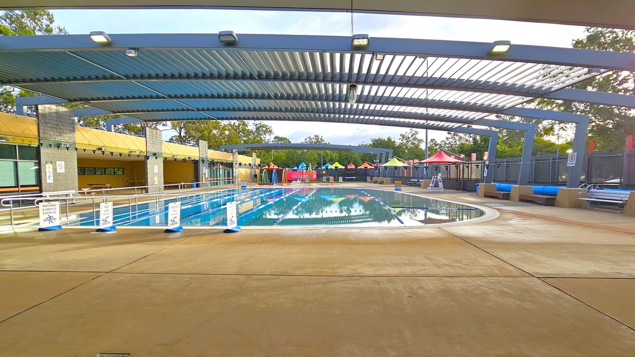 An outdoor swimming pool with clear water is surrounded by a concrete deck. Overhead, a metal pergola provides partial shade. On the left, there are signs indicating different swimming lanes and a covered area with tables. On the right, colourful umbrellas and seating areas are visible. In the background, trees and more seating areas can be seen. The pool is divided into lanes with floating markers.