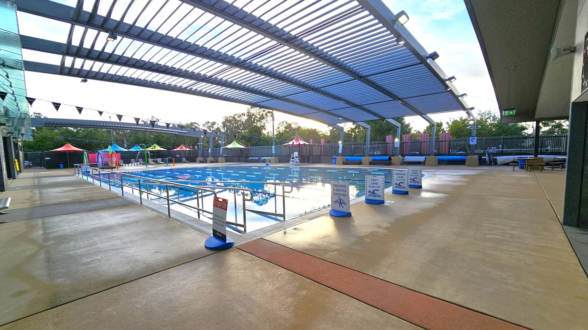 A covered swimming pool area with a slatted roof allowing some light through. The pool is in the centre, surrounded by signs indicating different lanes for walking, gentle exercise, and swimming speeds. On the left, there is a glass wall, and on the right, a solid wall with an exit sign. In the background, colourful tents and trees are visible. The floor is a mix of concrete and textured surfaces.