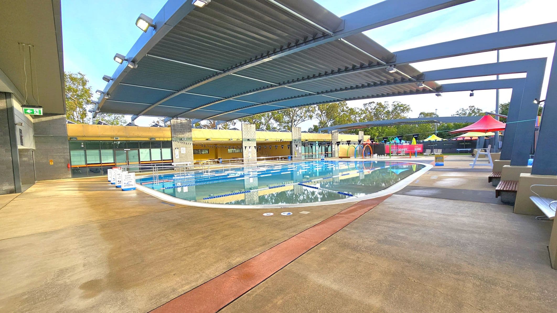 An outdoor swimming pool with clear water and lane dividers. A modern, curved metal roof structure provides partial shade. On the left, a concrete walkway with an exit sign. In the background, a yellow building with large windows and trees. On the right, colourful shade structures and seating areas. The floor is a mix of concrete and red pathways.