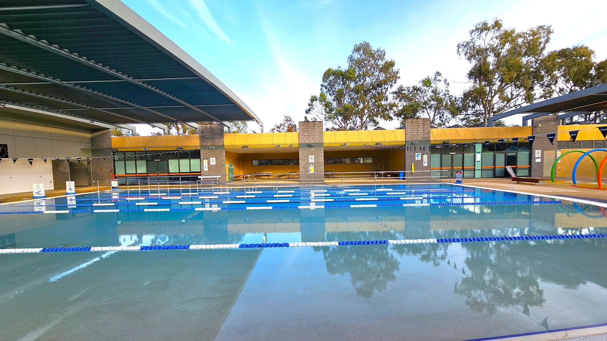 An outdoor swimming pool with blue and white lane dividers stretches across the foreground. The pool is partially covered by a modern, angular roof on the left. In the background, a yellow structure with large windows and brick columns houses seating and facilities. Trees are visible beyond the building. On the right, colourful arches are positioned near the poolside. The area is well-maintained and organised, with clear signage and safety features.