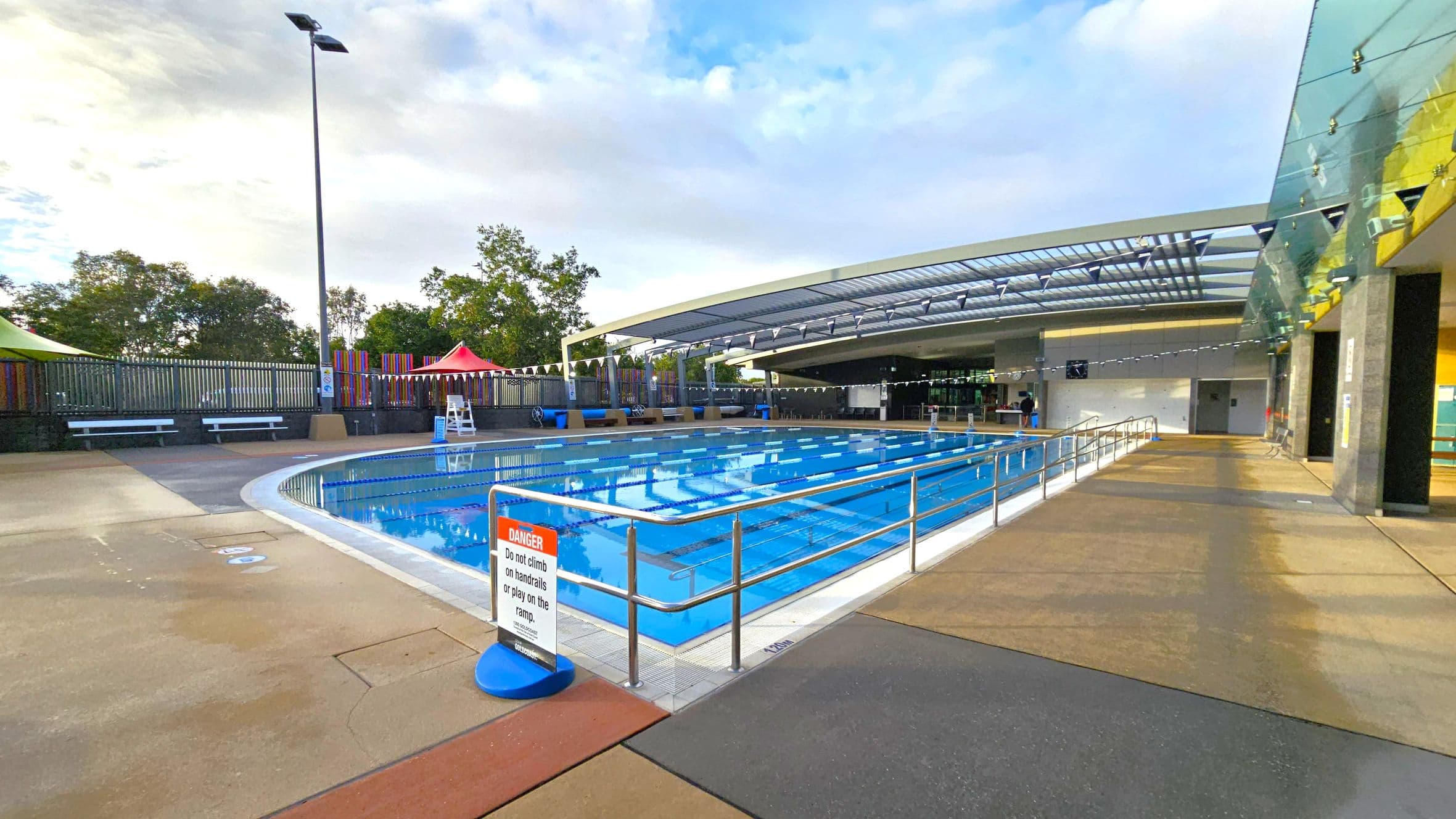 An outdoor swimming pool with clear blue water is in the centre, surrounded by a metal railing. A sign warns against climbing on the handrails. On the left, there are benches and a tall lamp post, with trees and colourful fencing in the background. On the right, a modern building with a glass roof and walls extends over part of the pool area. The floor is a mix of concrete and textured surfaces.