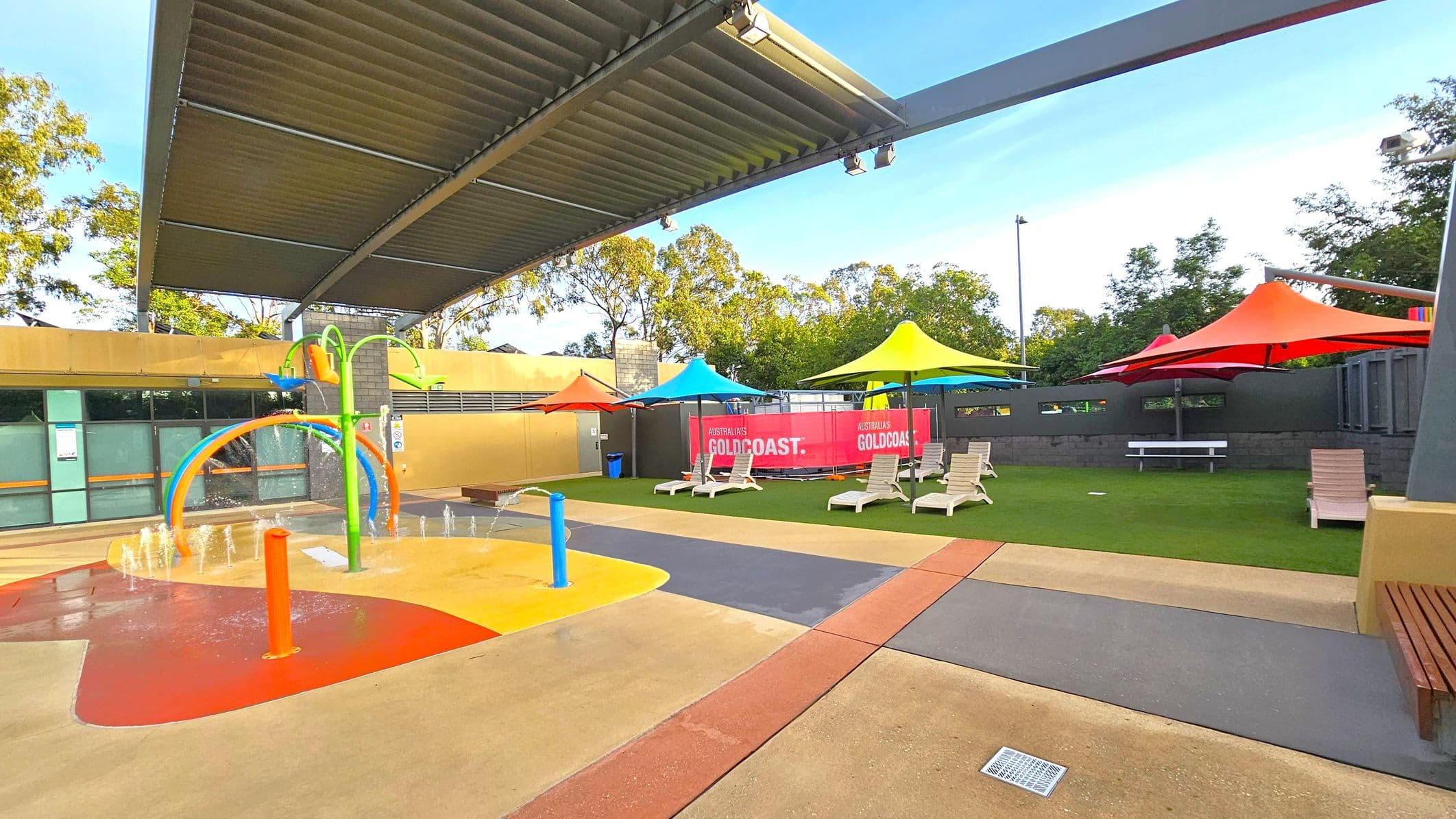 A children's splash pad with colourful play equipment is on the left, featuring water jets and a green arch with orange and blue elements. On the right, there are several sun loungers under vibrant yellow, blue, and red shade umbrellas on a grassy area. A pink banner with "GOLDCOAST" is visible in the background. The area is covered by a large, slatted metal roof. Trees are visible beyond the perimeter wall. The ground is a mix of textured concrete and rubber surfaces.
