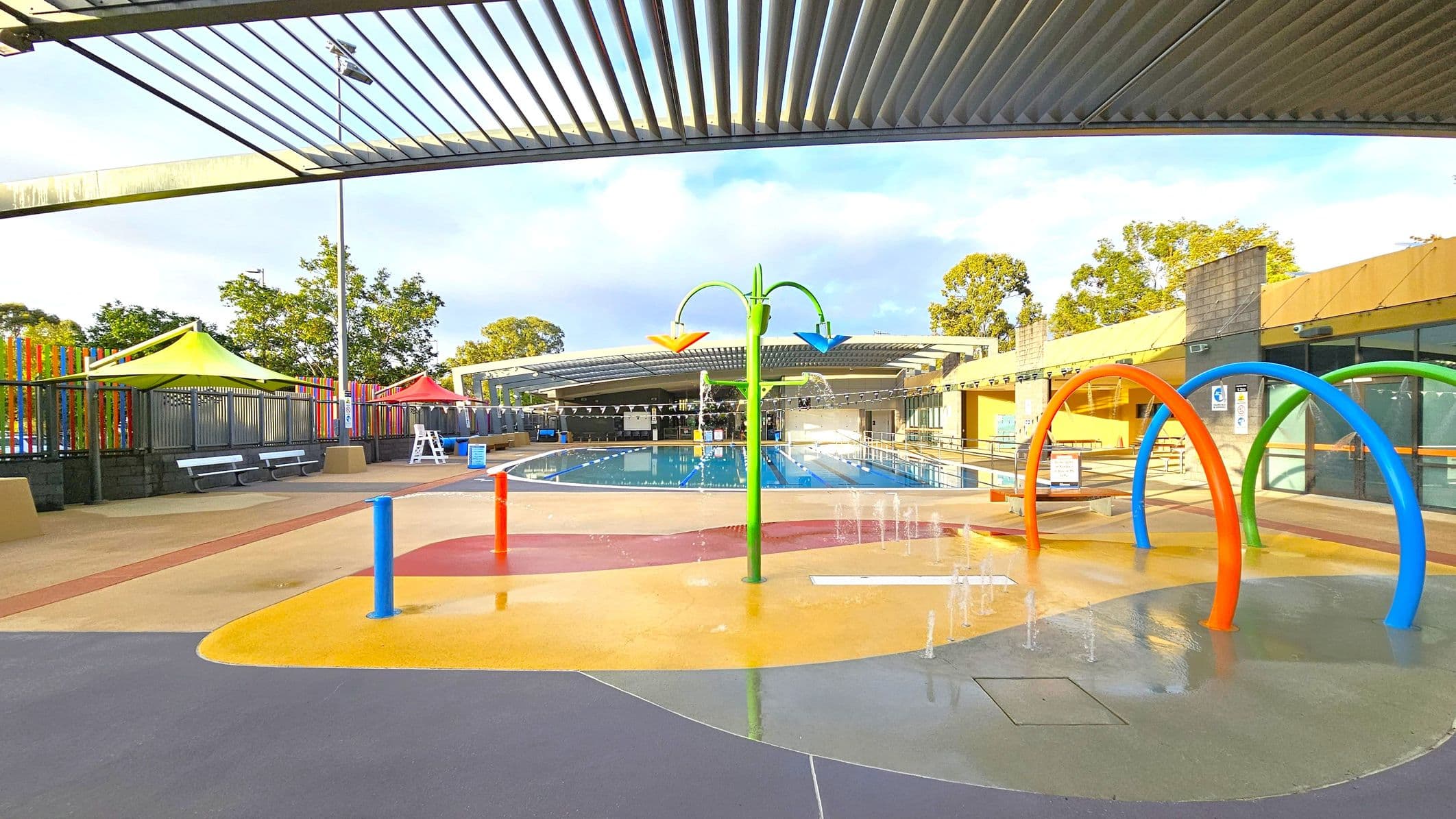 A colourful outdoor water play area with a splash pad in the foreground, featuring blue, orange, and green arches. Water sprays from the ground and a central green structure with orange and blue spouts. In the background, a swimming pool is visible, surrounded by a fence with multicoloured vertical slats. Yellow and red shade structures are on the left, with benches underneath. The ceiling has a slatted design, and trees are visible beyond the fence.