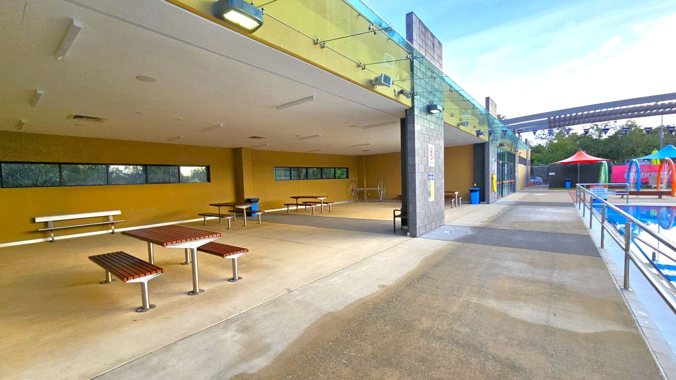 On the left, there is a covered area with yellow walls, featuring several picnic tables and benches. The ceiling is white with lights. In the middle, a grey pillar separates the covered area from the open walkway. On the right, a pathway runs alongside a swimming pool with a metal railing. In the background, colourful tents and a structure with a slatted roof are visible. The floor is a mix of concrete and textured surfaces.