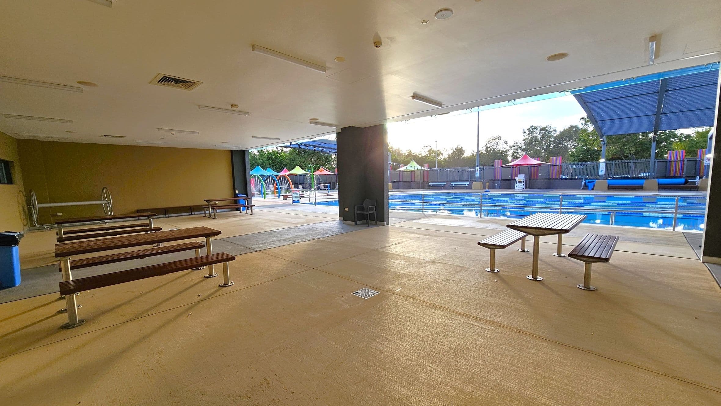 An indoor area with a beige floor and ceiling, featuring several wooden benches and tables. To the right, a swimming pool with blue lanes is visible, partially covered by a large canopy. In the background, colourful structures and a fence line the pool area. Trees are visible beyond the fence.