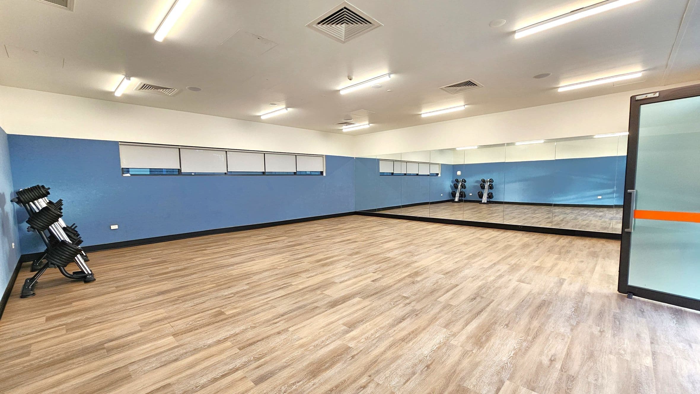A spacious room with wooden flooring and a blue accent wall. On the left, there is a rack of stacked weights. The right side features a large mirror covering the wall, reflecting the room's interior. A glass door with an orange stripe is partially visible on the right. The ceiling is equipped with fluorescent lights and air vents.