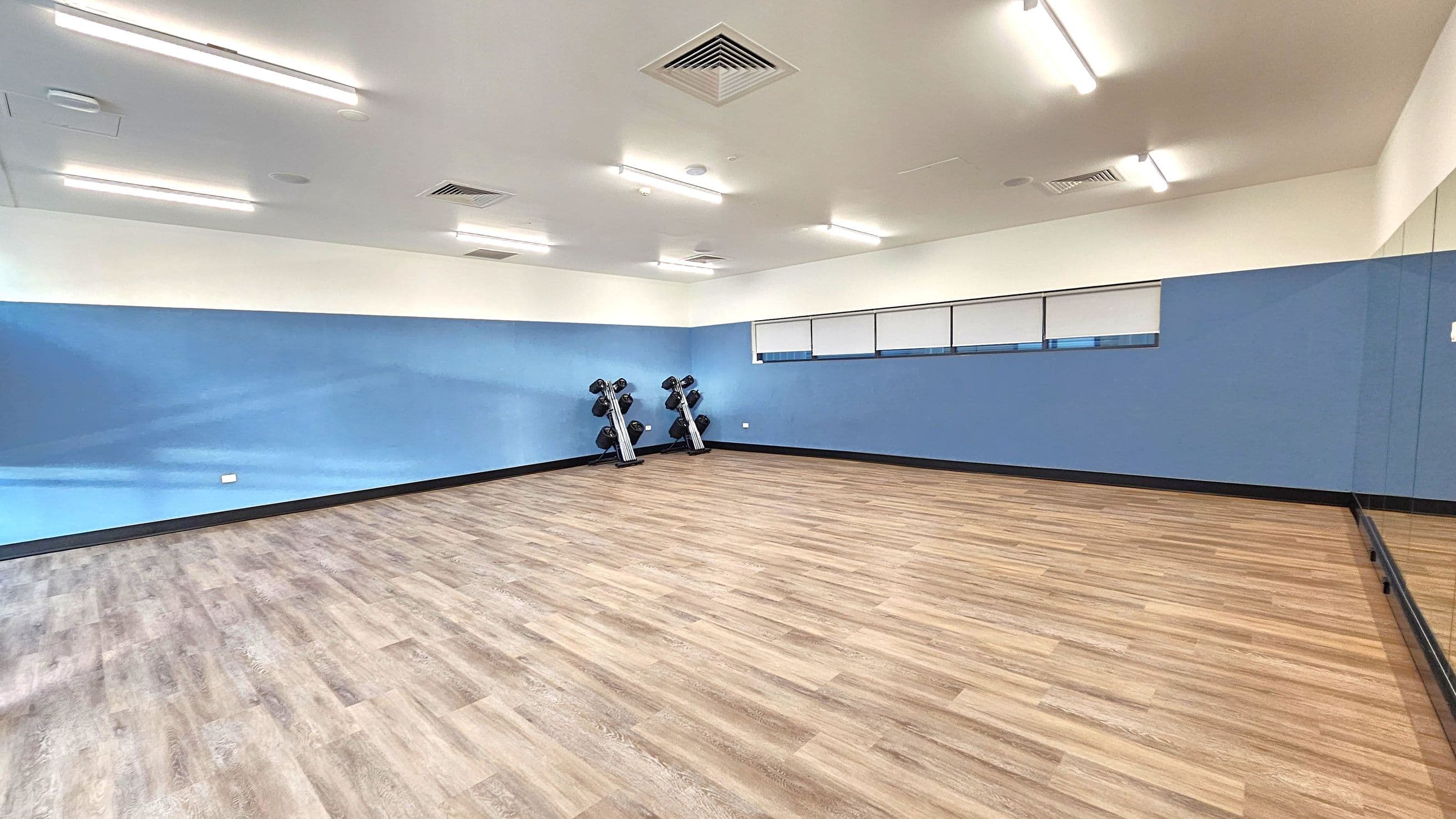 A spacious room with light wooden flooring and blue walls. On the left, there are two sets of dumbbells on racks against the wall. The right side features a large mirror extending along the wall. The ceiling is white with several fluorescent lights and air vents. The room has a clean and minimalistic appearance.