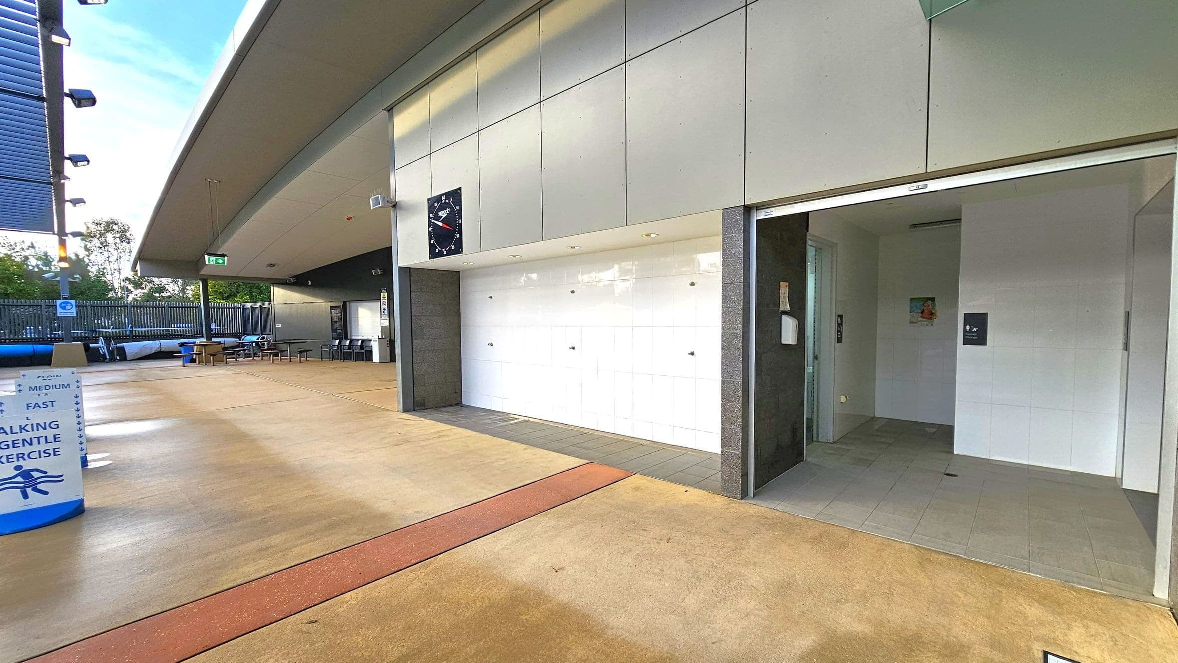 A modern outdoor area with a covered walkway. On the right, there's an entrance to a facility with white tiled walls and a sign. In the middle, a large clock is mounted on the wall above a white tiled section. On the left, there are tables and chairs under a roof, with a fence and trees in the background. The floor is a mix of light brown and red tiles, and the ceiling is a light grey with lighting fixtures. A sign on the left indicates walking and exercise lanes.
