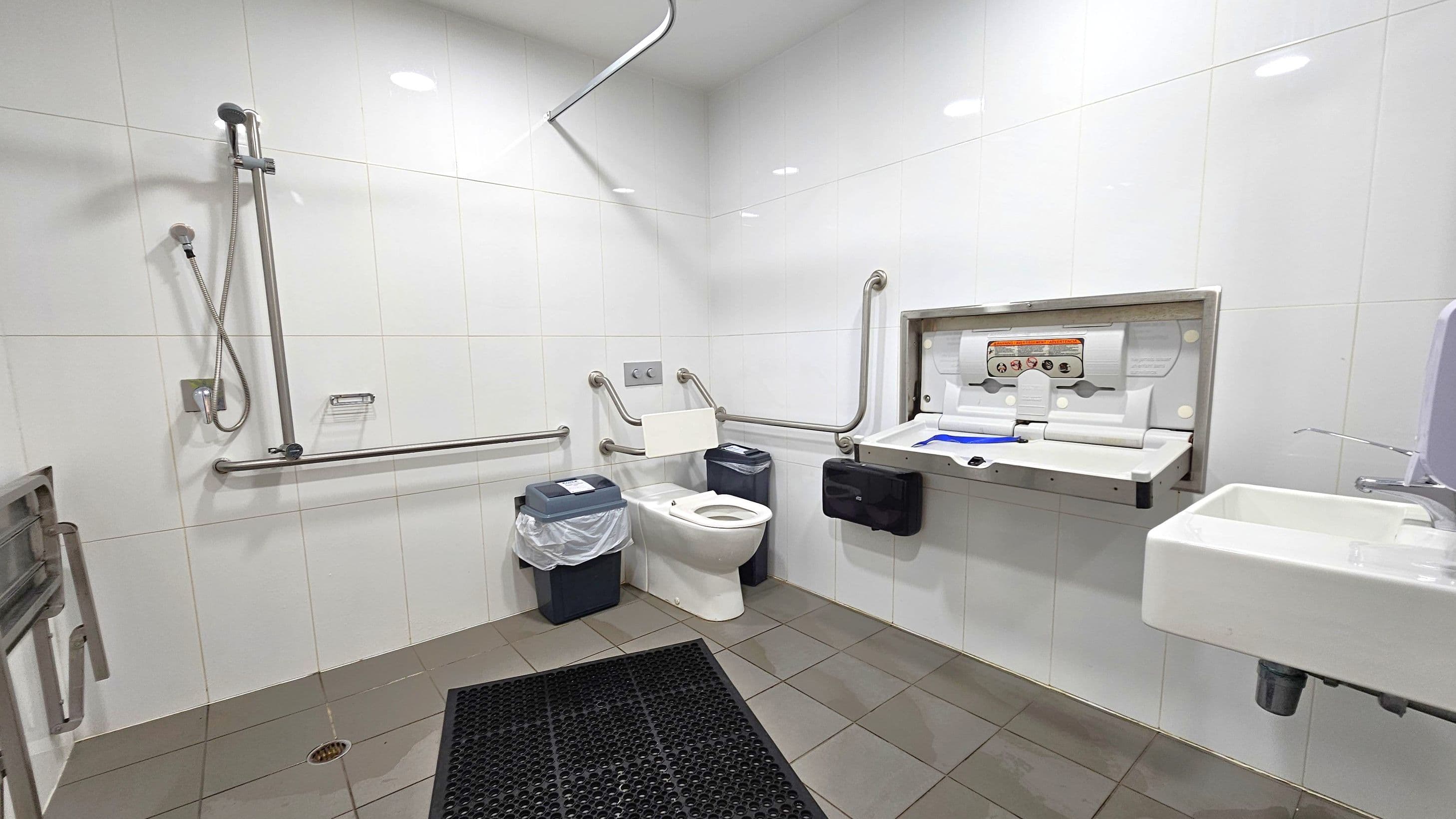 A spacious, accessible bathroom with white tiled walls and a grey-tiled floor. On the left, a shower with a handheld showerhead and grab bars. In the centre, a toilet with support rails and a bin with a liner. On the right, a baby changing station is mounted on the wall above a black paper towel dispenser. A small white sink with a soap dispenser is also on the right. The ceiling is white with recessed lighting.