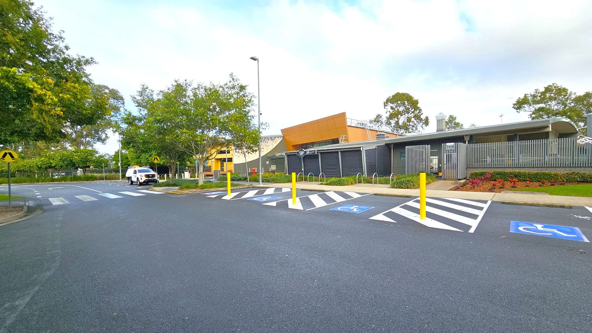 A car park with designated disabled parking spaces marked in blue and white is in the foreground. Yellow bollards separate the spaces. On the right, a modern building with grey and orange panels is visible, surrounded by landscaped gardens. In the background, a white vehicle is parked near a pedestrian crossing. Trees with green foliage line the left side, and a lamppost stands near the centre. The road surface is smooth and dark grey.