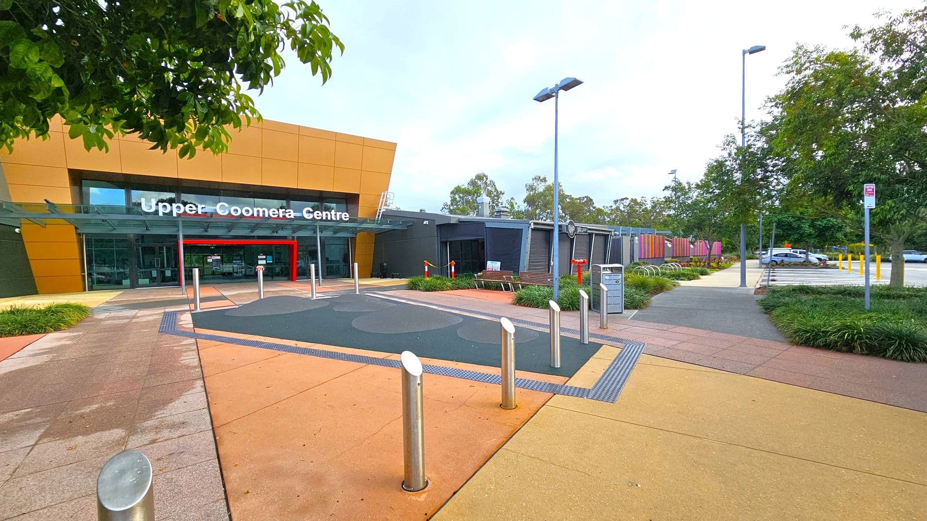A modern building with a large sign reading "Upper Coomera Centre" above the entrance. The facade is a mix of orange and grey panels. In front, there are several metal bollards and a pathway with various coloured tiles. To the right, a pathway leads to a car park with several parked cars. Trees and greenery are visible on both sides, with a few benches and a bicycle rack near the entrance. The area is well-lit with tall street lamps.