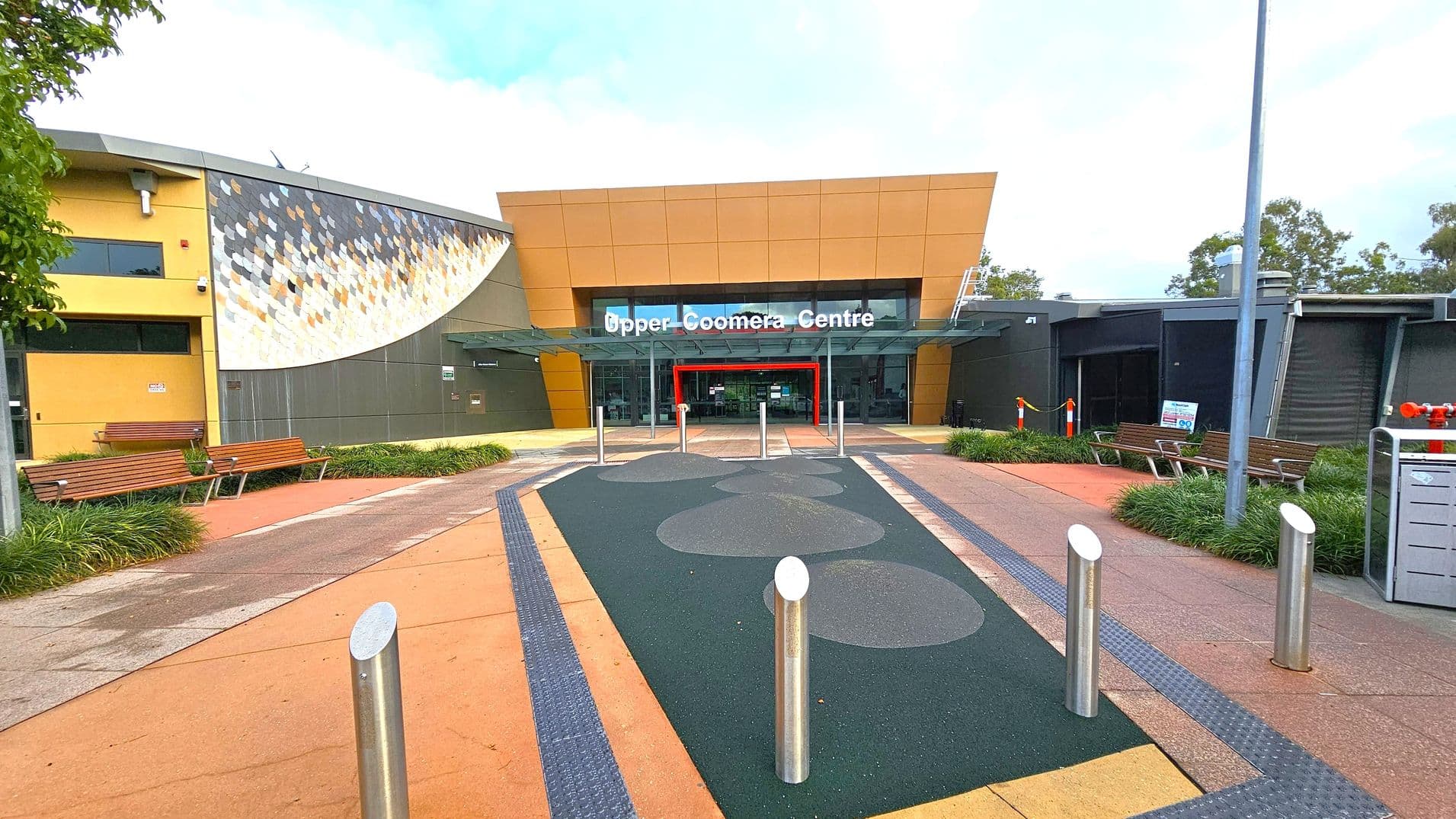 A modern building entrance with a sign reading "Upper Coomera Centre" above glass doors. The structure features a mix of yellow and grey panels, with a decorative mosaic on the left. Benches and greenery line the pathway leading to the entrance. The ground is paved with a combination of orange and dark green surfaces, and several metal bollards are positioned near the entrance. A tall lamp post is visible on the right.