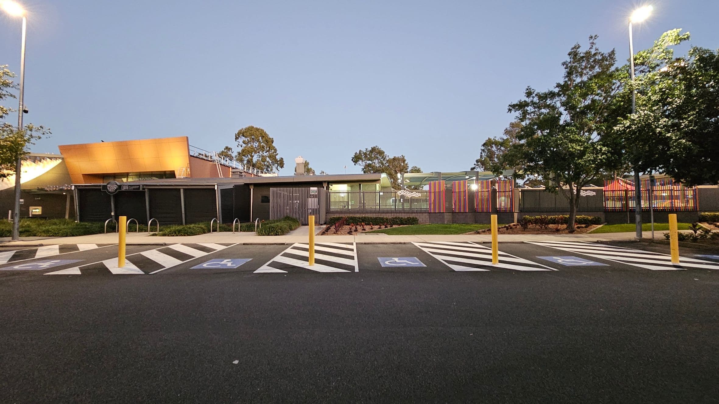 A parking area with marked disabled spaces in the foreground, featuring blue and white symbols. Yellow bollards separate the spaces. In the background, a modern building with a golden-brown facade and a section with colourful vertical slats. Trees are on both sides, and streetlights illuminate the scene. The ground is asphalt, and the area is well-maintained with some greenery.