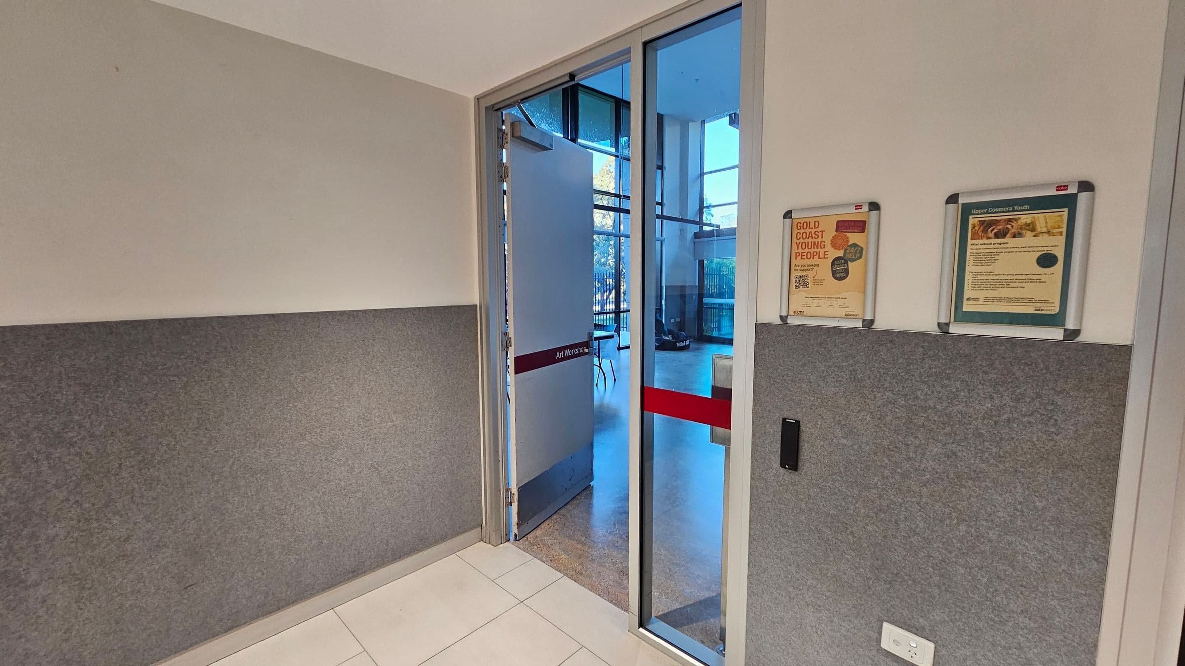 A hallway with grey and beige walls. On the right, two framed notices are mounted on the wall. The floor is tiled in white. A glass door with a red stripe, labelled "Art Workshop," is partially open, leading to a room with large windows and visible tables and chairs. An electrical outlet is near the bottom of the right wall.