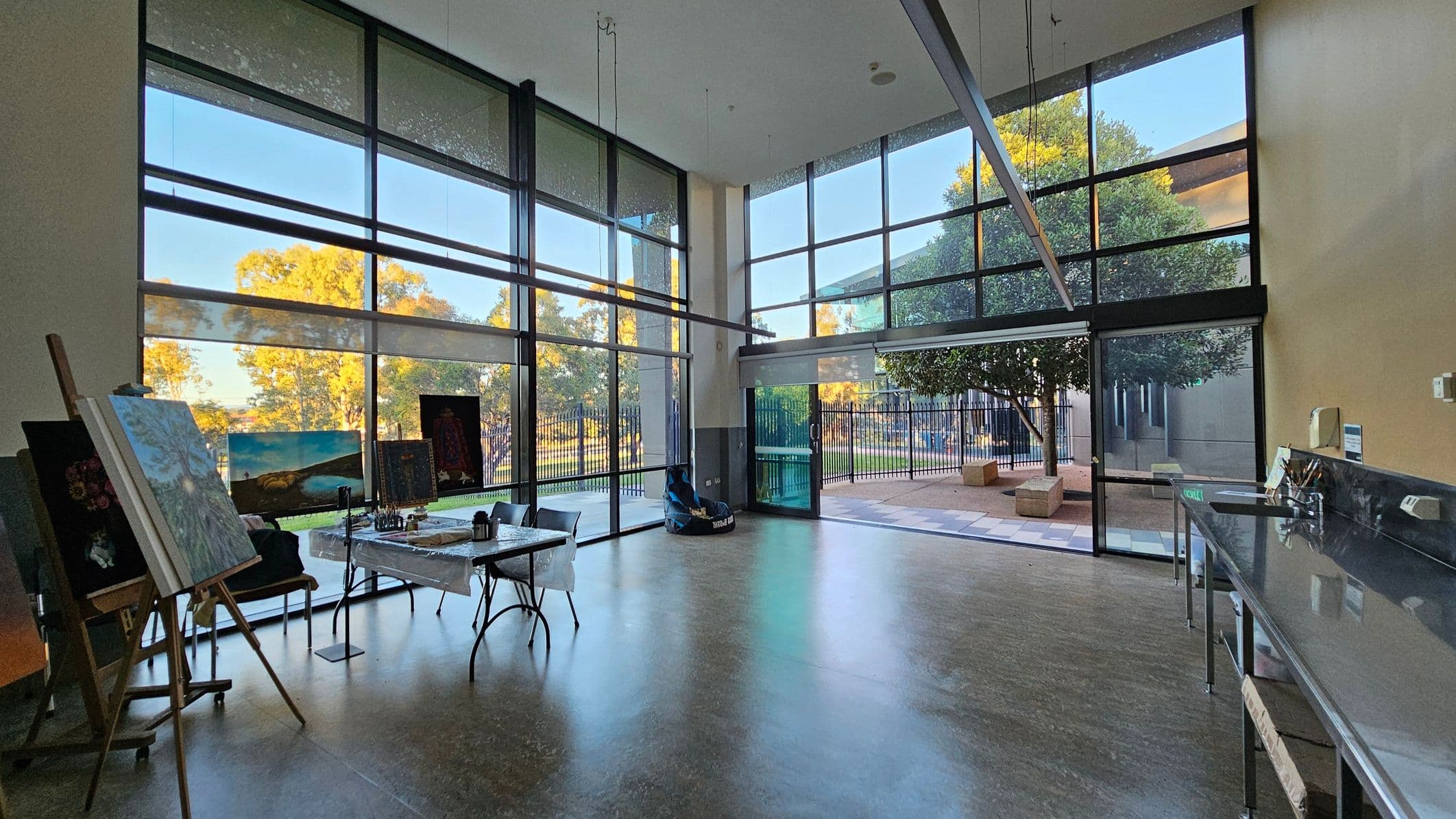 A spacious art studio with large floor-to-ceiling windows on the left and centre, allowing natural light to flood in. On the left, several easels display paintings, and tables hold art supplies. The floor is a smooth, light-coloured surface. On the right, a long counter with a sink is visible. Outside, through the windows, there are trees and a patio area with benches. The ceiling is high, enhancing the open feel of the space.