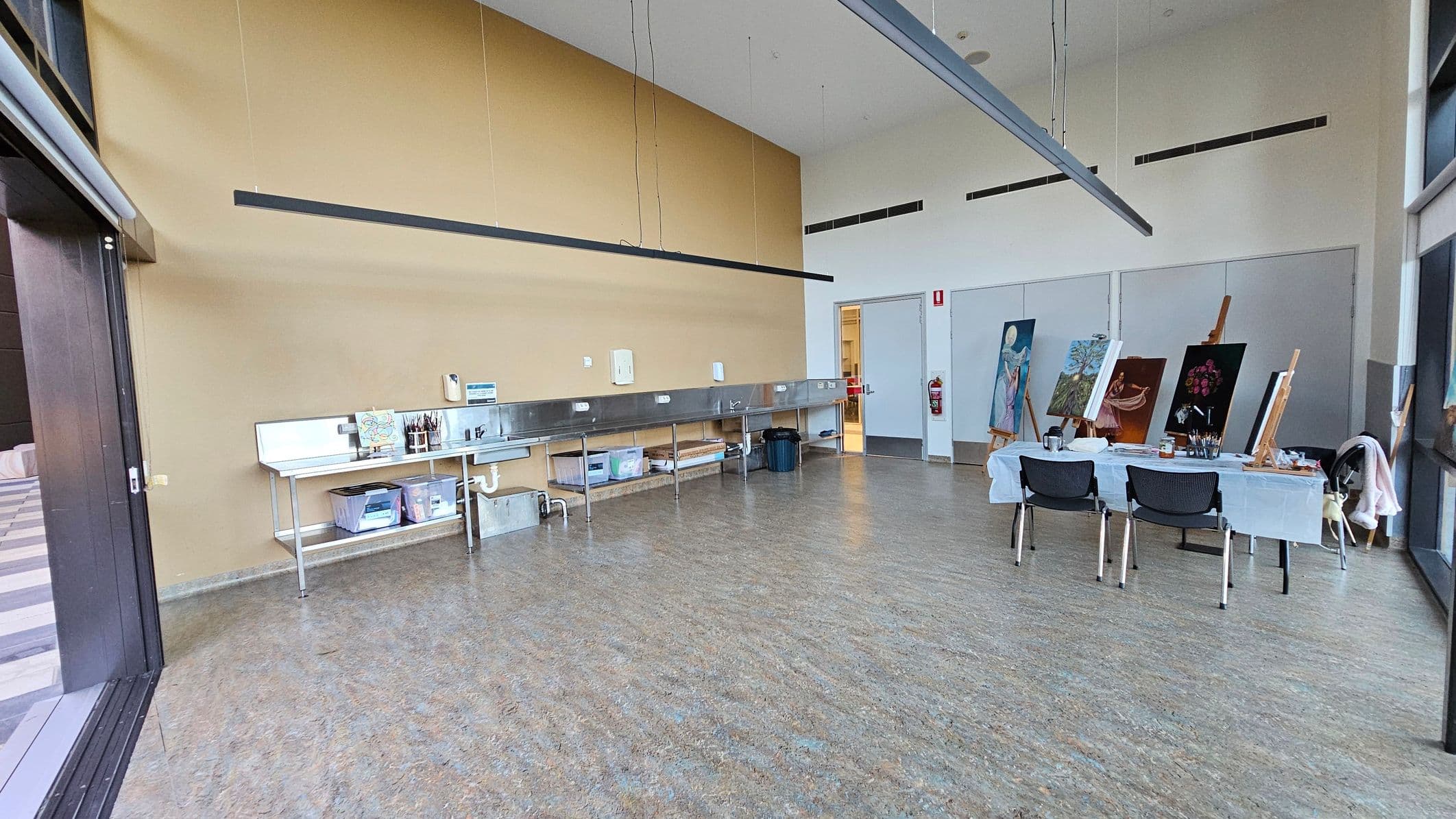 A spacious art studio with a light brown wall on the left featuring a long metal sink and storage shelves underneath. Various art supplies are visible on the shelves. On the right, a table covered with a white cloth holds several easels displaying paintings. Two chairs are positioned in front of the table. The floor is speckled grey, and the ceiling has long, suspended light fixtures. Large windows on the right allow natural light to fill the room.