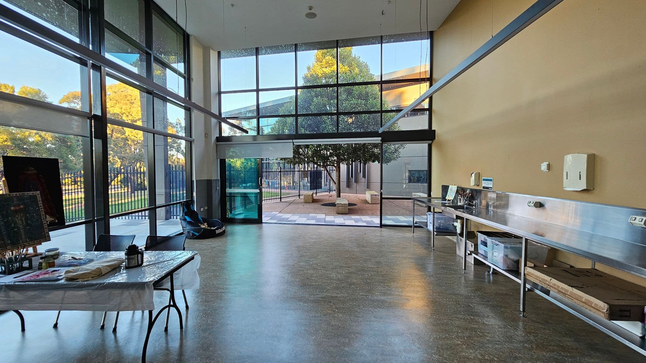 A spacious room with large floor-to-ceiling windows on the left and centre, offering a view of trees and an outdoor area. A table with art supplies and a canvas is on the left. The right side features a long stainless steel counter with storage boxes underneath. The floor is a polished, light-coloured surface, and the ceiling is high, enhancing the open feel of the space.