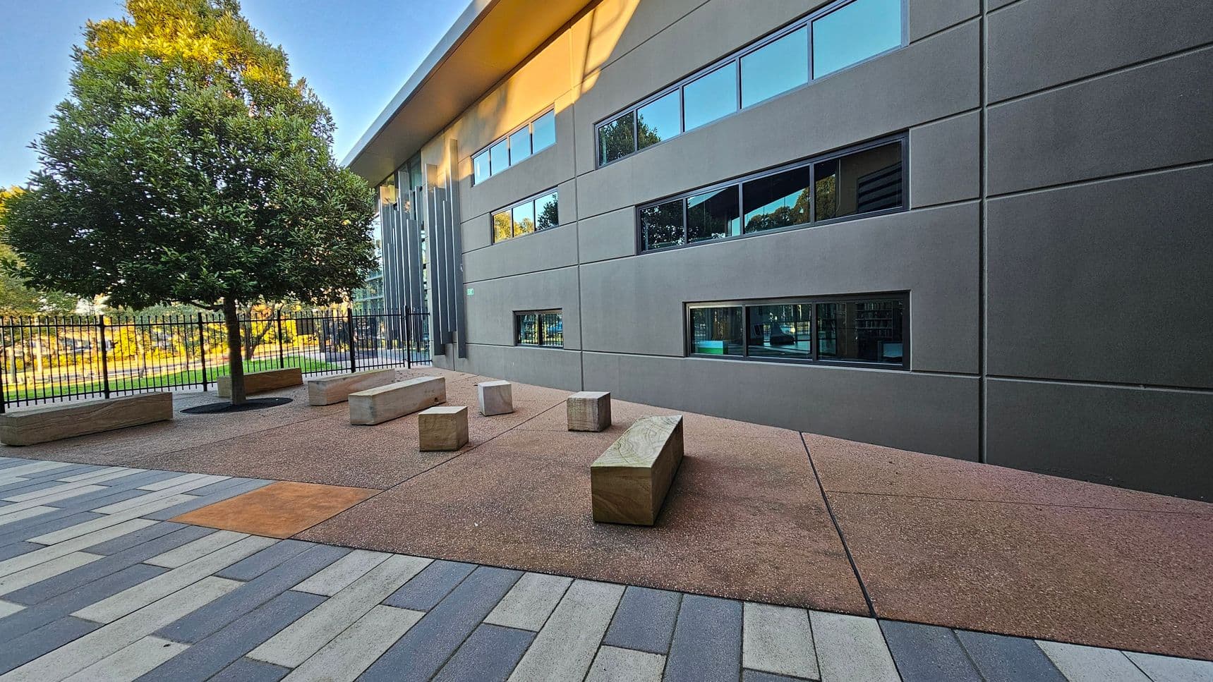 A modern outdoor area with a large tree on the left, surrounded by a black metal fence. The ground is a mix of textured brown and grey paving stones. Several rectangular stone benches are scattered around. On the right, a building with a smooth grey facade features several horizontal windows. The building's roof has a slight overhang, casting shadows on the wall.
