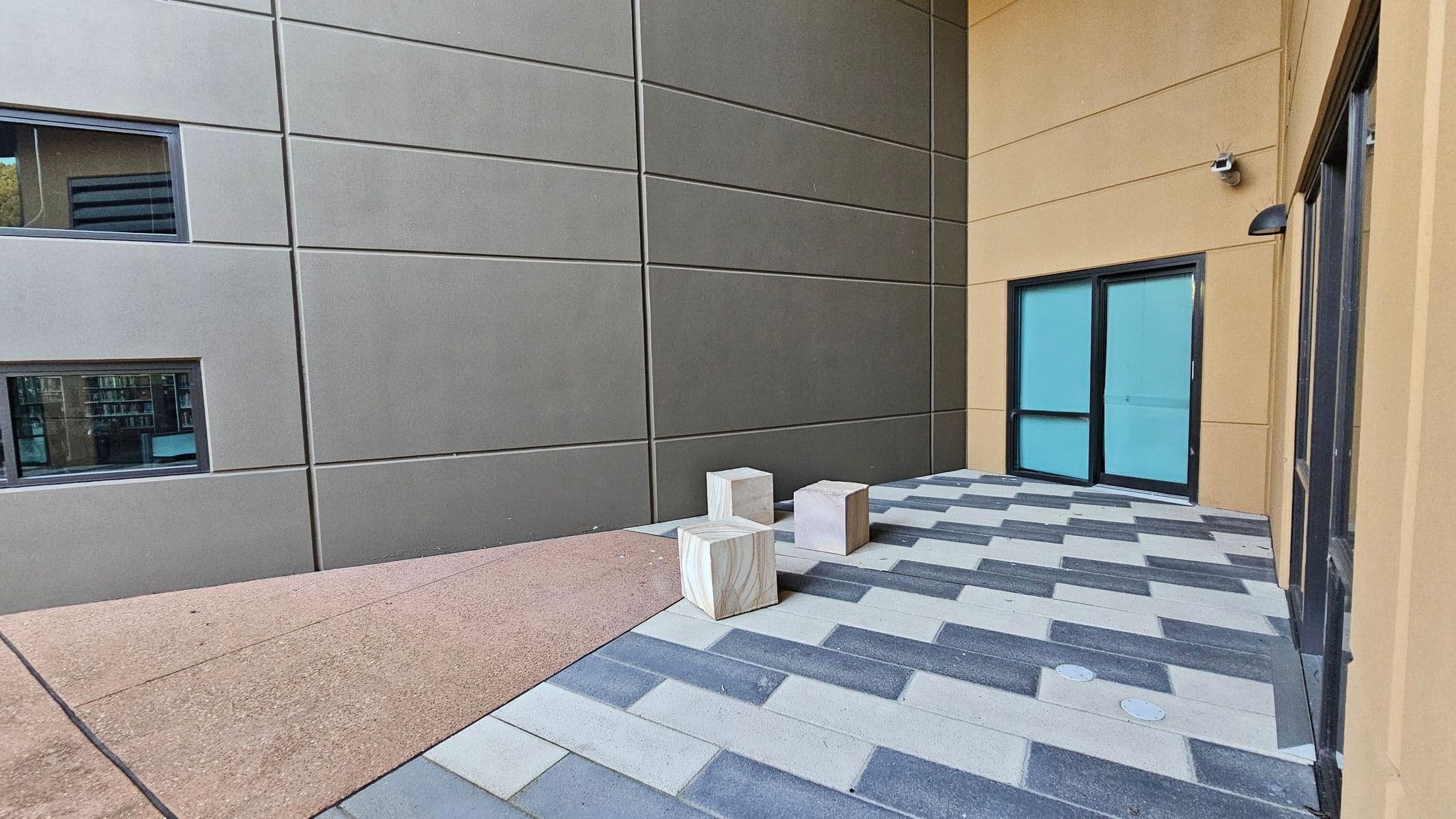A small outdoor area with a tiled floor featuring a pattern of grey and beige rectangles. On the right, there are two large windows with blue-tinted glass set in a beige wall. A security camera is mounted above the windows. On the left, a grey wall with two narrow horizontal windows. Three cube-shaped stone stools are placed on the tiled floor. The ground transitions from tiles to a textured beige surface.