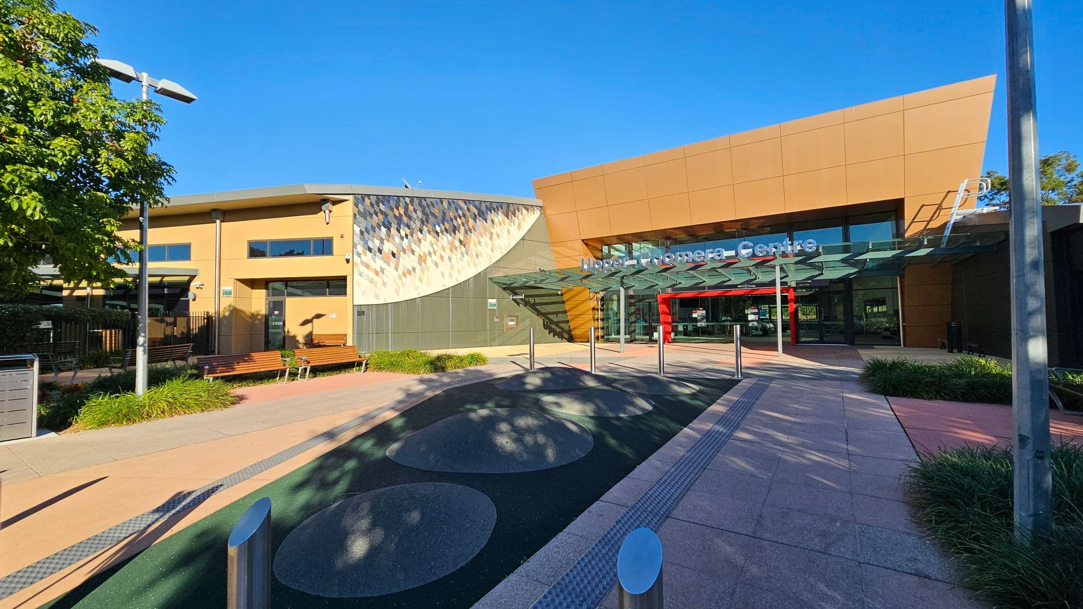 A modern building with a glass entrance is on the right, featuring a sign above the door. The structure has a geometric design with a mix of tan and grey panels. On the left, there is a smaller building with a mosaic wall and several windows. Benches and greenery line the walkway, which has circular patterns on the ground. A tall lamp post and a tree are visible on the left, and the area is well-lit by sunlight.