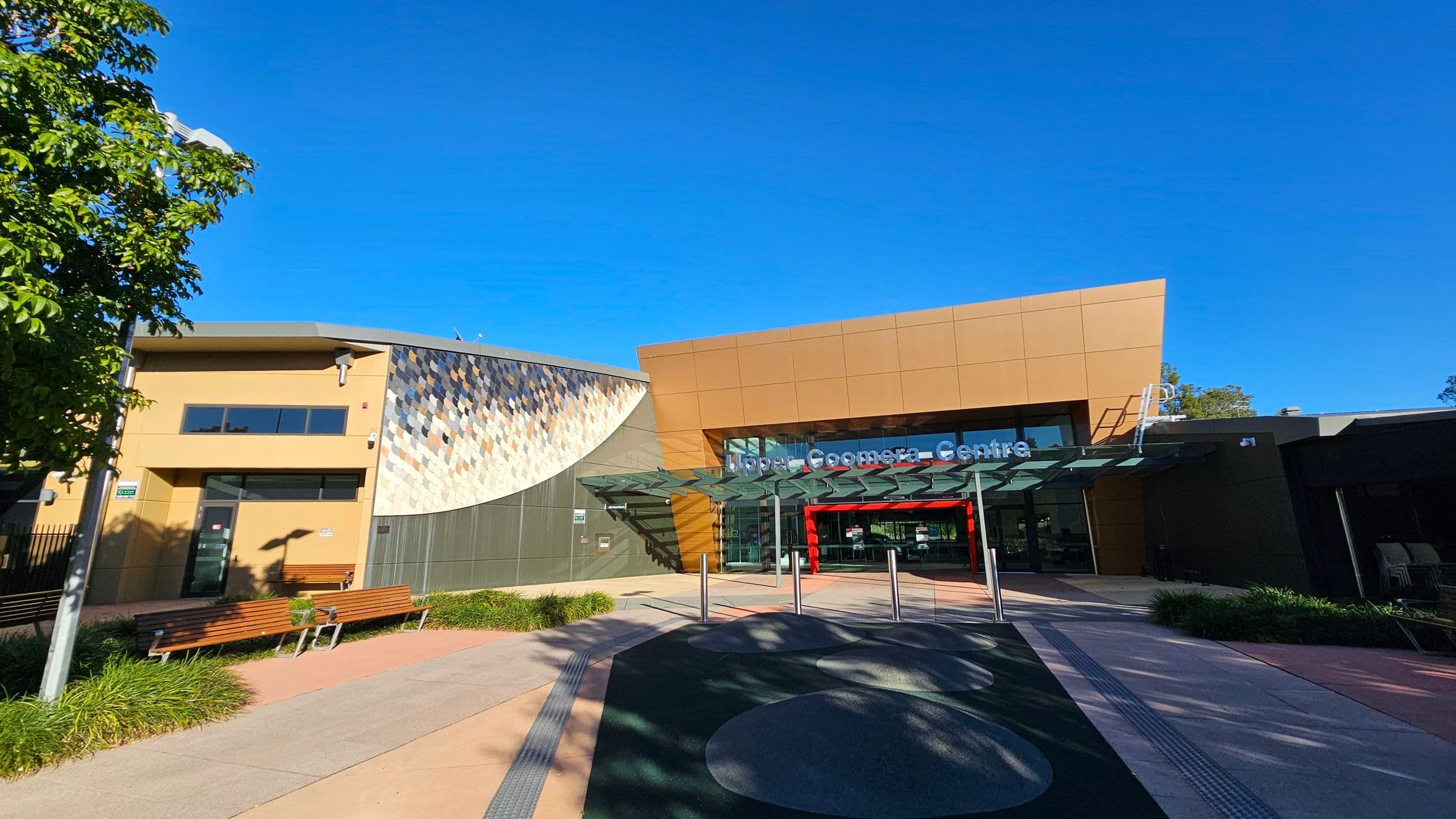 A modern building with a glass entrance is in the centre, featuring a sign that reads "Upper Coomera Centre." The structure has a mix of tan and grey panels, with a mosaic pattern on the left side. In the foreground, there are benches and landscaped greenery. A tree is visible on the left, casting shadows on the pavement. The sky is clear and blue.