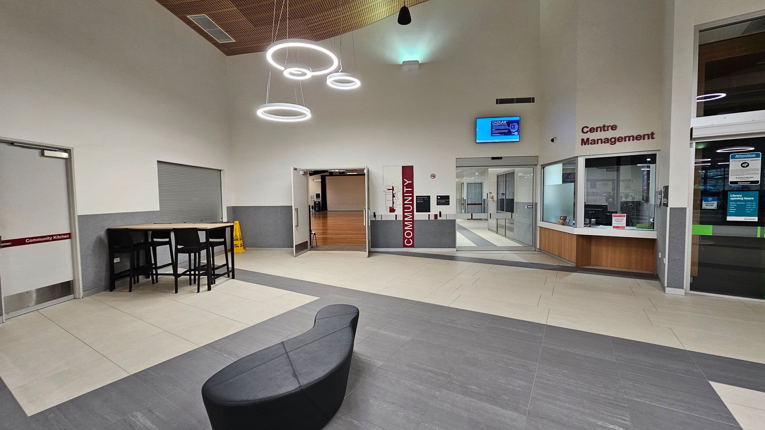 A modern interior space with a grey and beige tiled floor. On the left, there's a door labelled "Community Kitchen" and a high table with black chairs. In the centre, double doors lead to a room with wooden flooring, marked "Community". Above, circular light fixtures hang from the ceiling. On the right, a "Centre Management" office with a glass window and counter. A digital screen is mounted above the office. A curved black bench is in the foreground.