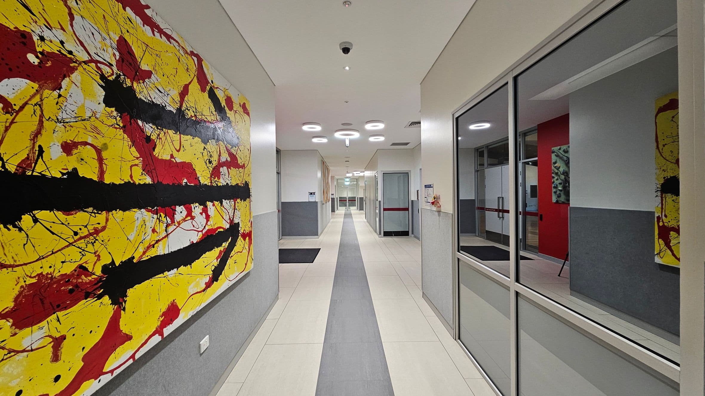 A hallway with a large abstract painting on the left wall, featuring bold black, red, and yellow splashes. The floor is tiled in light colours with a central grey strip. On the right, there are glass panels reflecting the painting and part of a red wall. The ceiling is fitted with circular lights and a security camera. The corridor extends into the distance with doors and more artwork visible at the far end.