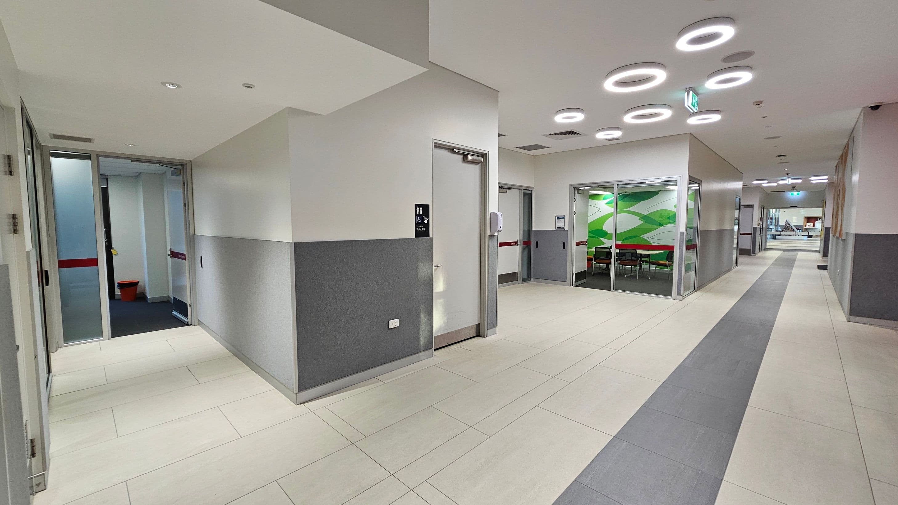 A modern hallway with light-coloured tiled flooring and grey walls. On the left, there are doors with frosted glass panels. In the centre, a door with a restroom sign and a wall-mounted hand sanitiser. To the right, a glass-walled room with a green abstract design on the wall and visible chairs inside. The ceiling features circular light fixtures. The hallway extends into the distance with more doors and a visible exit sign.