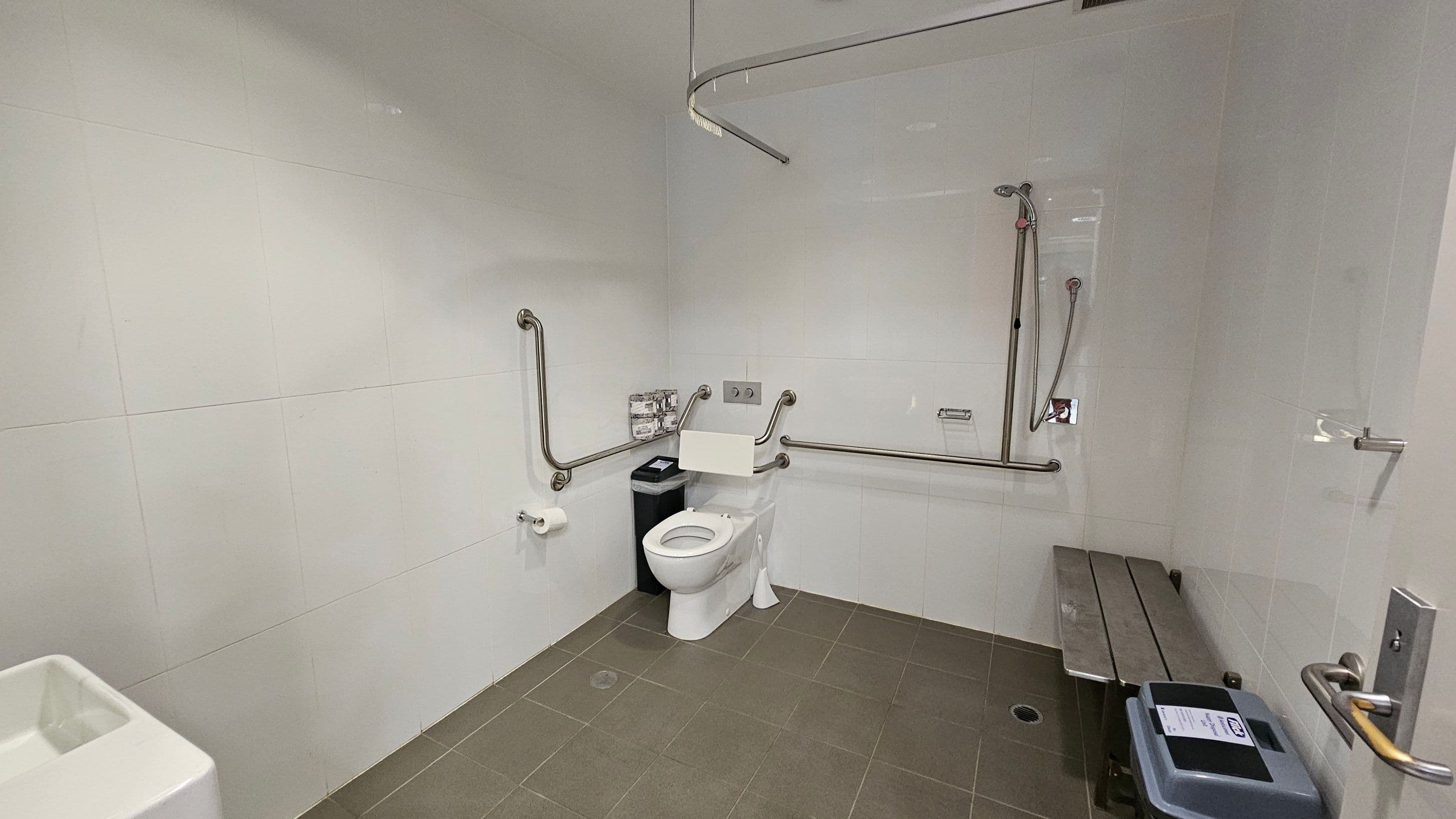 A spacious accessible bathroom with white tiled walls and a green-tiled floor. In the centre, there is a toilet with support rails on both sides. To the right, a shower area with a handheld showerhead and additional grab bars. A bench is positioned against the right wall. On the left, a toilet paper holder and a small bin are visible. The ceiling has a curved rail, possibly for a shower curtain.