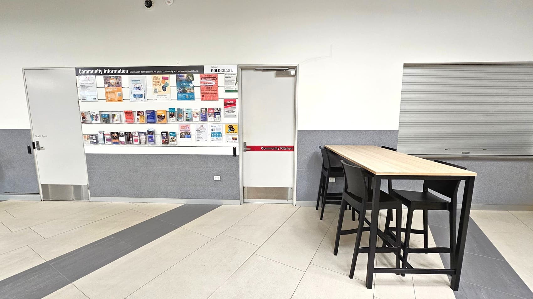 On the left, a community information board is filled with various pamphlets and notices. Below it, a door marked "Staff Only" is visible. In the centre, a door labelled "Community Kitchen" is present. On the right, a long wooden table with several black chairs is positioned in front of a closed metal shutter. The floor is tiled in shades of grey and beige, and the walls are painted white with a grey lower section.